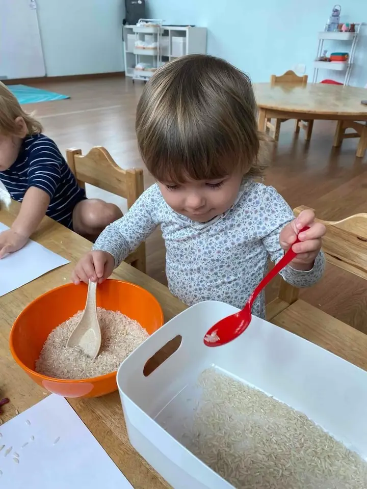 A toddler spooning rice between two bowls at a classroom table while another child works beside them