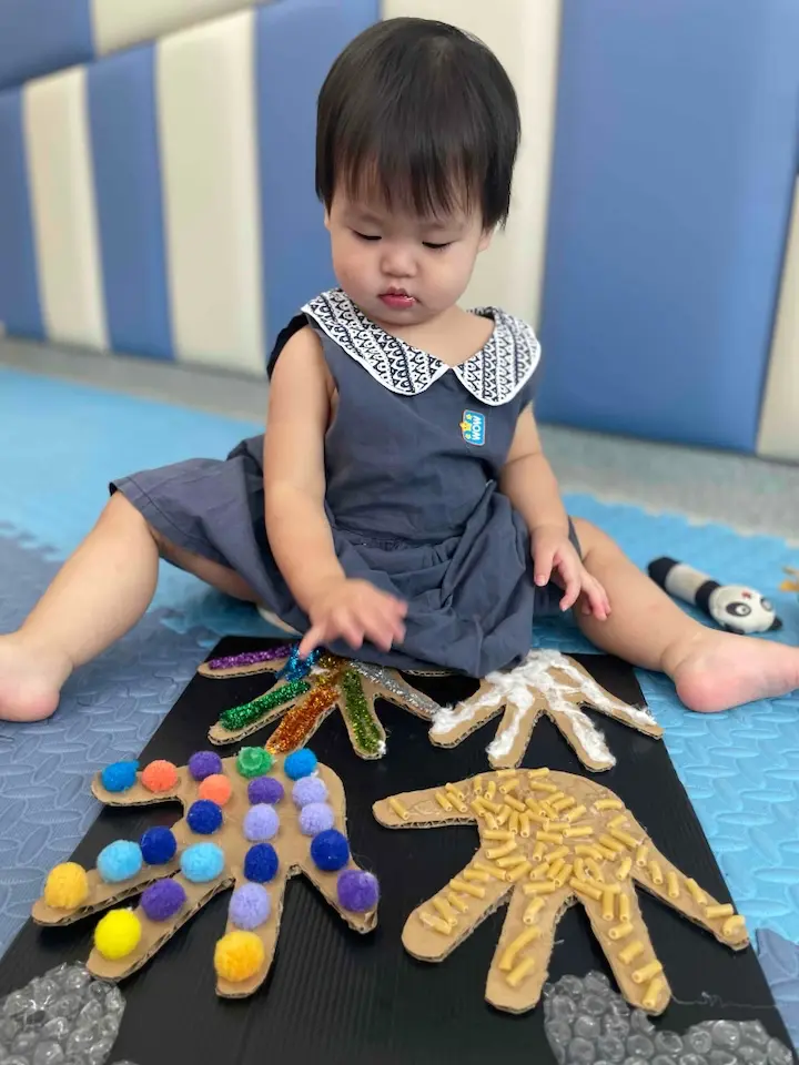 A toddler concentrating on a textured handprint craft on a classroom floor